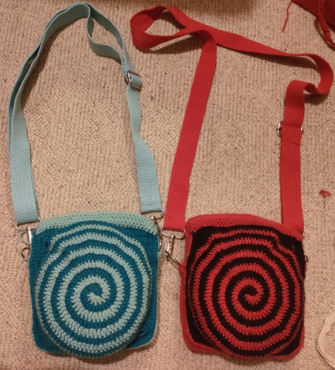 two crochet satchels with a swirly circular front pocket, left is turquoise and teal, right is black and red.