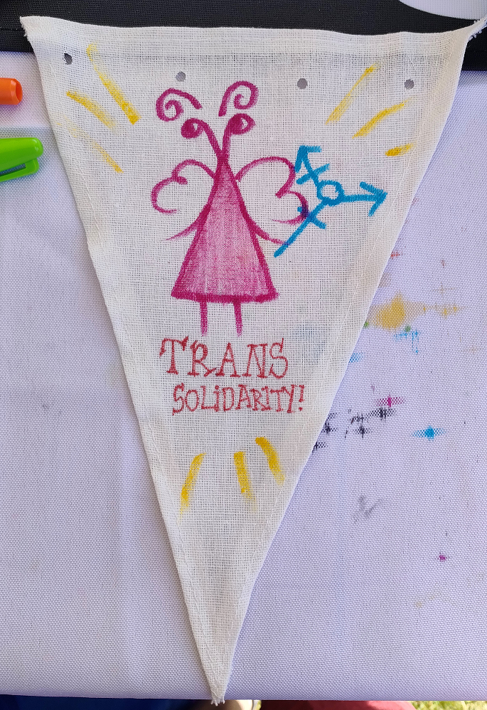 One white bunting triangle with an insectlike pink fairy holding a blue trans symbol. It says 'trans solidarity!' and there are yellow lines coming off it.