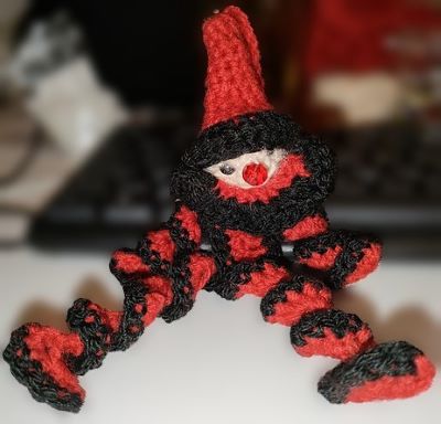 Small crocheted red and black clown with spiral limbs, a red nose and a pointy hat sitting somewhat folornly. 