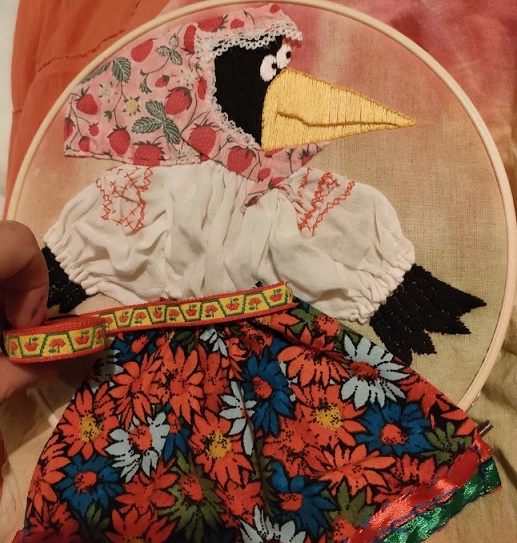 applique and embroidery of smiling cartoon crow wearing flowery red skirt and white blouse with a babushka-headscarf.