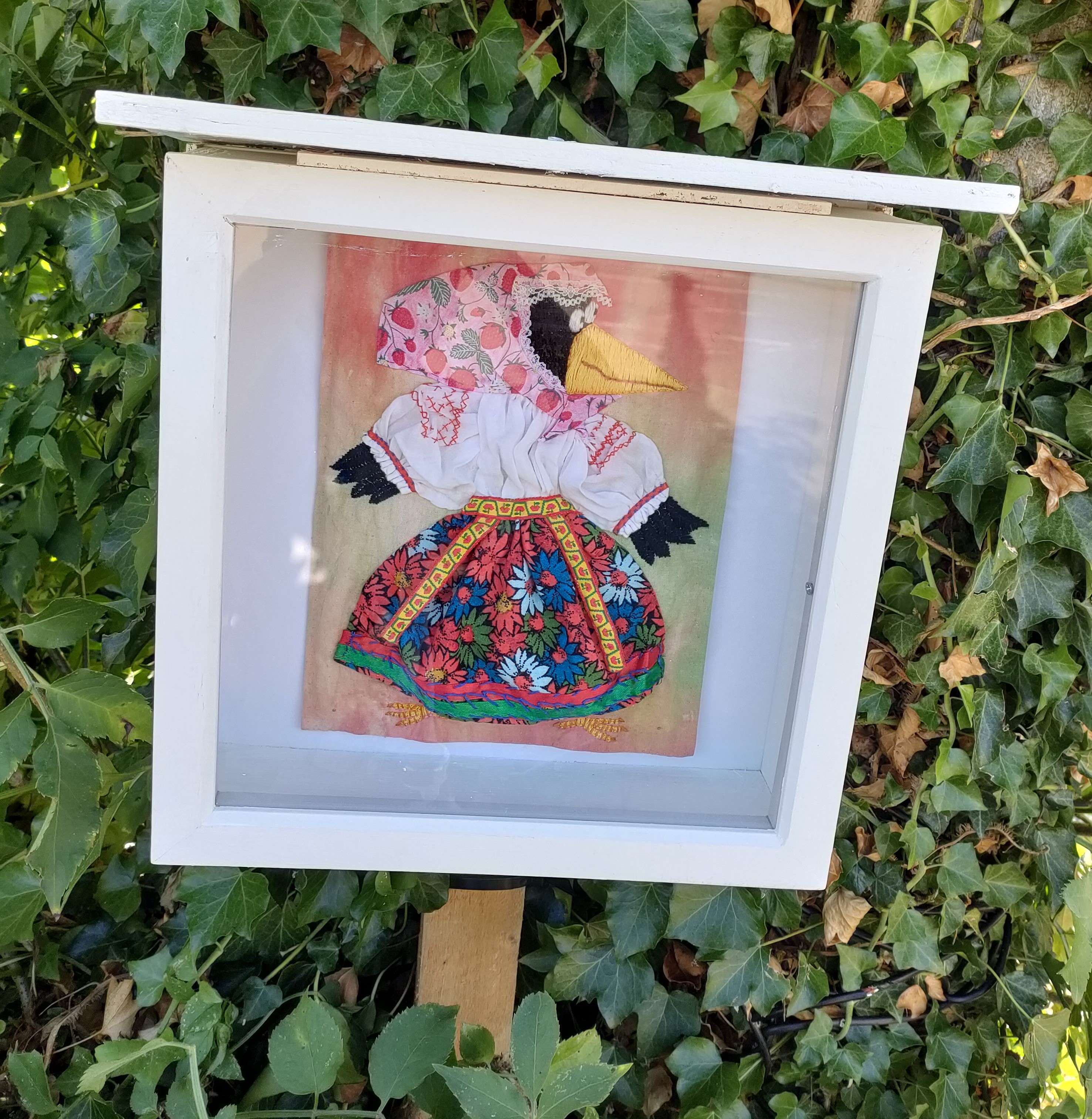 cartoon-style crow wearing folk dress embroidery framed displayed outdoors.